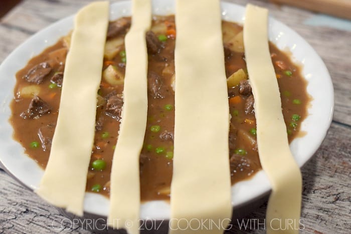 Beef pot pie filling in a pie dish topped with four strips of pie crust dough.