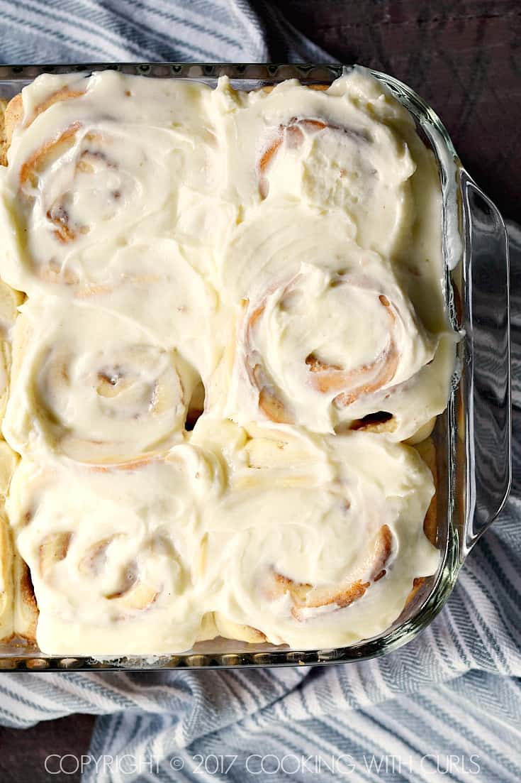 A thick layer of cream cheese icing spread over the top of cinnamon rolls in a baking dish.