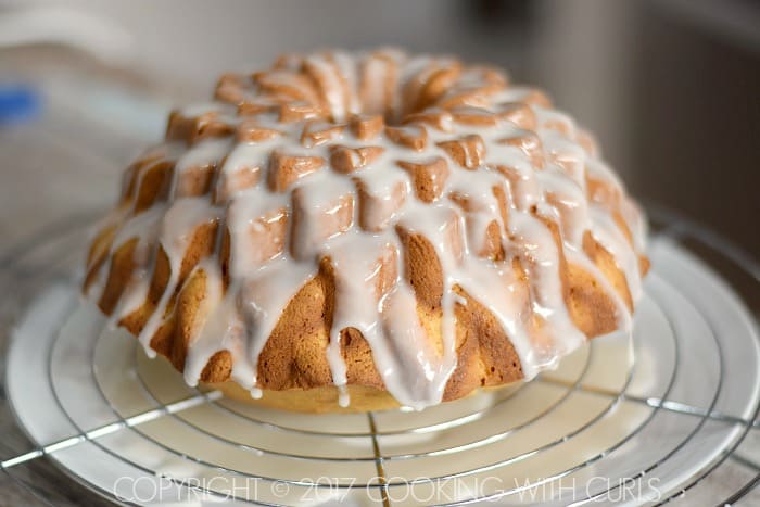 Glaze dripping down the sides of a flower shaped bundt cake onto a round cooling rack on a large plate.