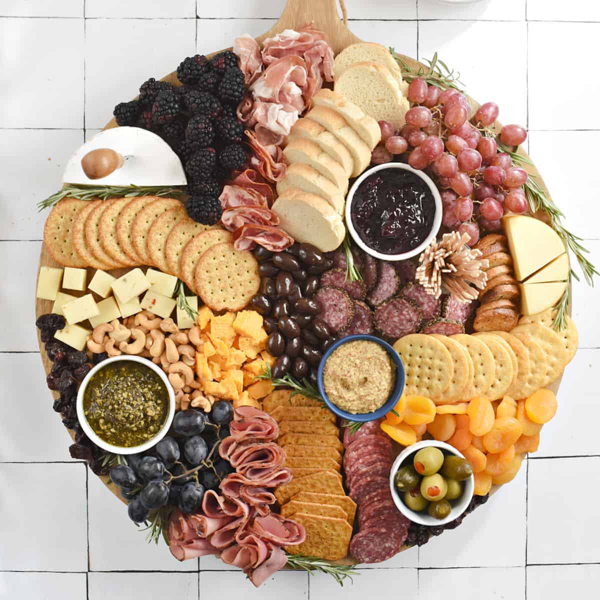 A large round board topped with slices of meats, cheeses, bread, crackers, dried apricots and cherries, grapes, and nuts.