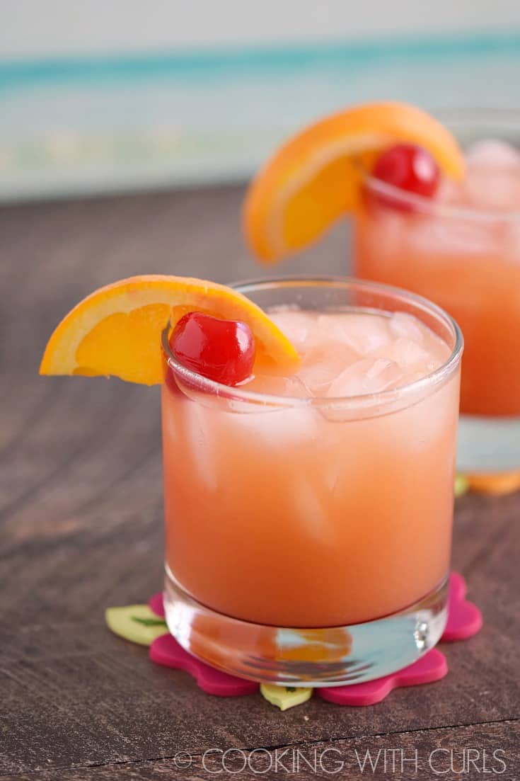 Looking down at two short glasses filled with a bright orange-pink drink garnished with orange wedges and a cherry.