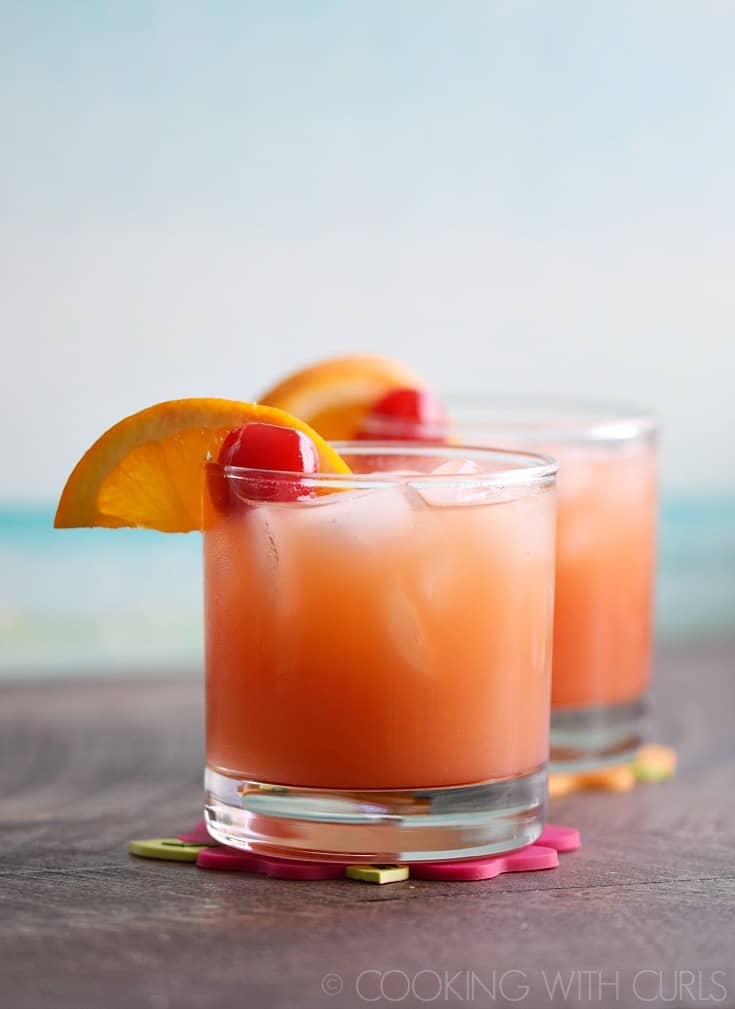Two short glasses filled with a bright orange-pink drink garnished with orange wedges and a cherry.