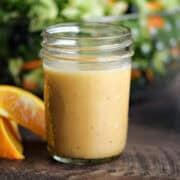 Homemade citrus vinaigrette salad dressing in a glass jar next to orange slices and a bowl of salad.