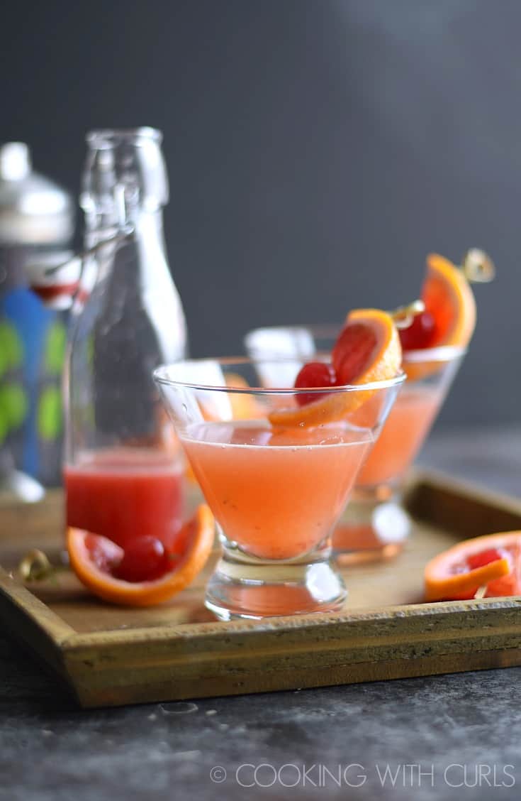 Two Blood Orange Martinis on a wood tray garnished with an orange slice and cherry.