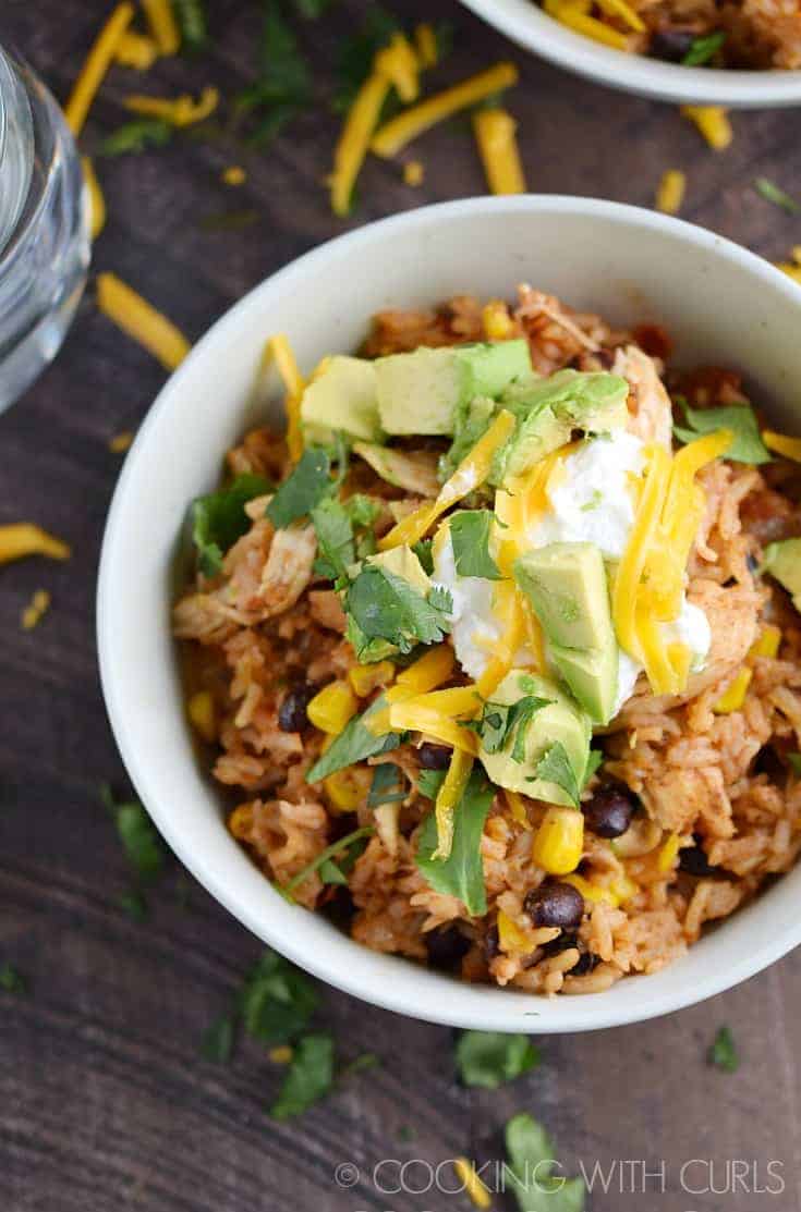 Chicken, rice, corn, black beans diced avocado, sour cream and shredded cheddar cheese in a white bowl.