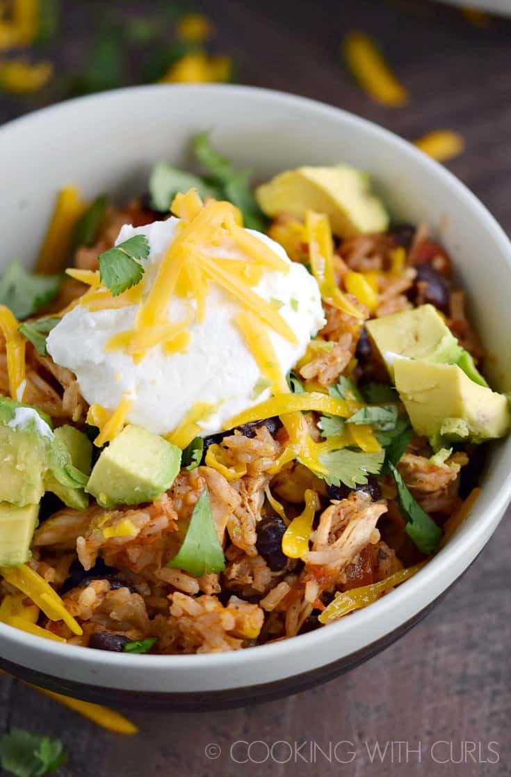 Taco chicken, diced avocado, rice and black beans topped with sour cream and shredded cheddar cheese in a white bowl.