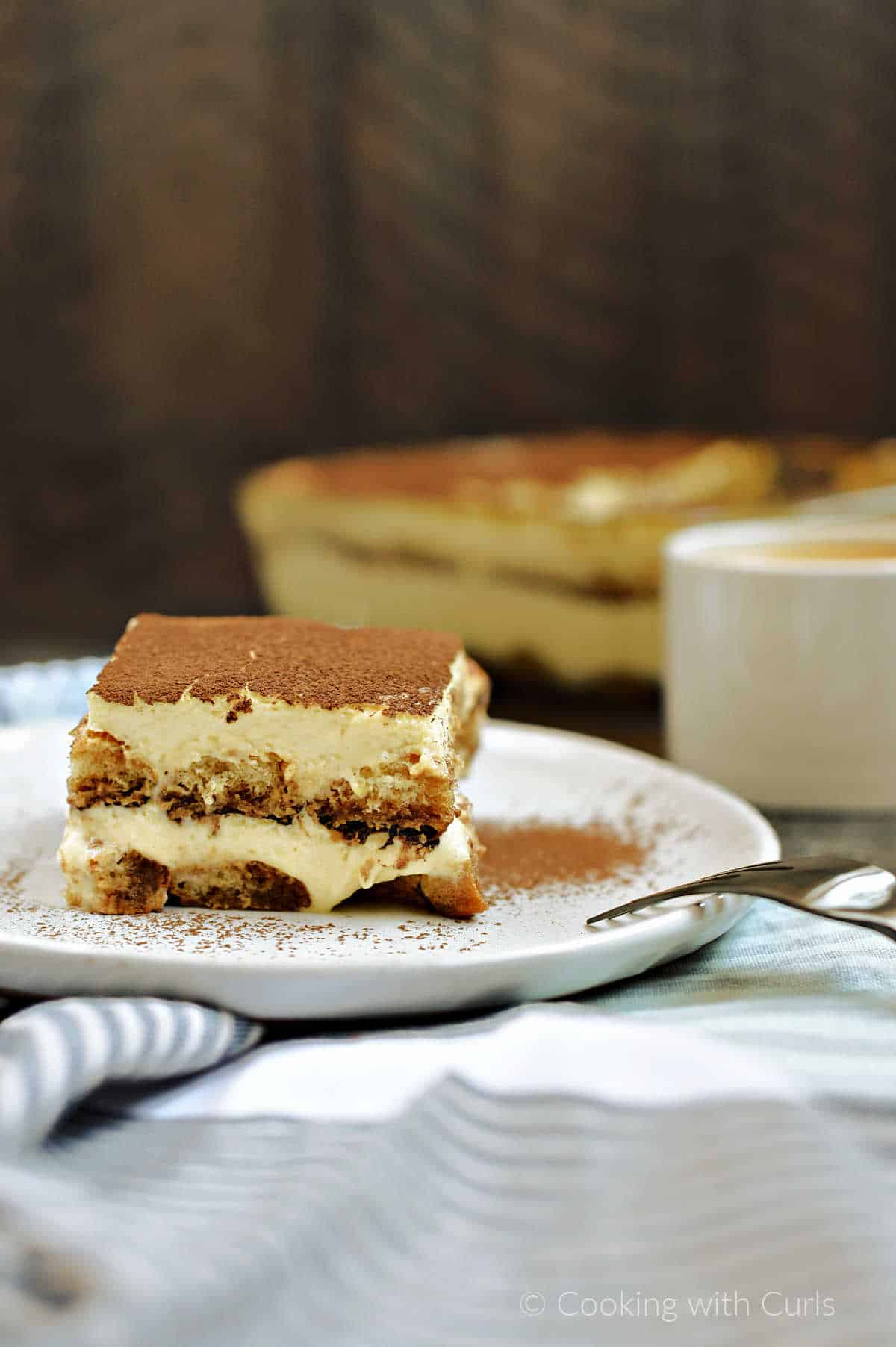 A slice of layered tiramisu on a plate with a pan of tiramisu and cup of coffee in the background.