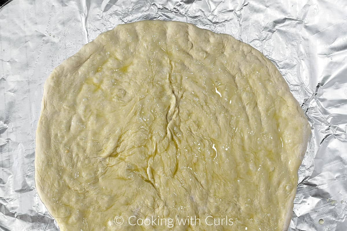 Flattened pizza dough on a sheet of foil brushed with oil. 