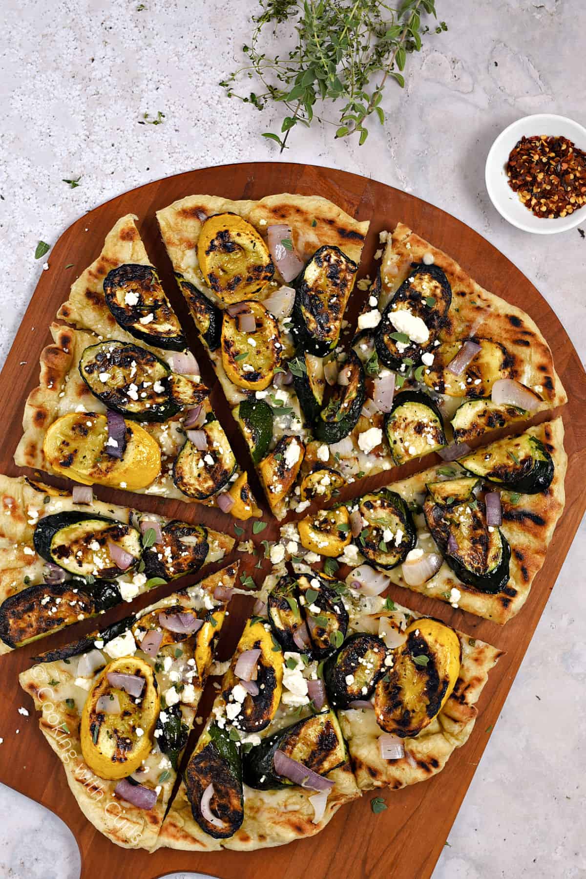 Looking down on a grilled pizza topped with summer squash, red onion, cheese and fresh herbs on a pizza peel.