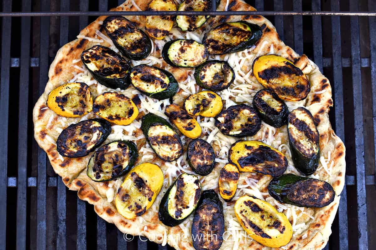 Grilled pizza crust topped with grated mozzarella cheese and grilled squash on a gas grill. 