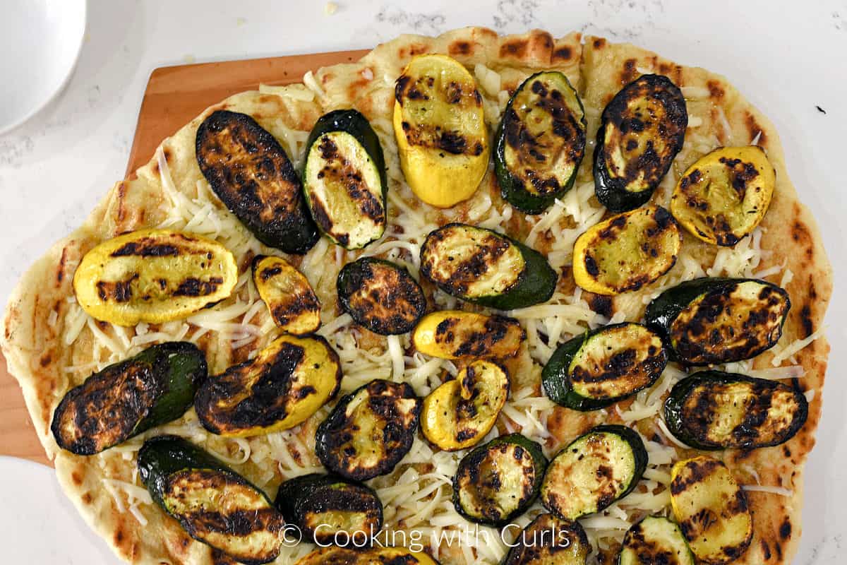 Grilled pizza crust topped with grated mozzarella cheese and grilled vegetables on a pizza peel.