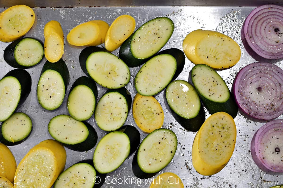 Sliced zucchini, yellow squash and red onion brushed with oil and sprinkled with salt and pepper on a baking tray.