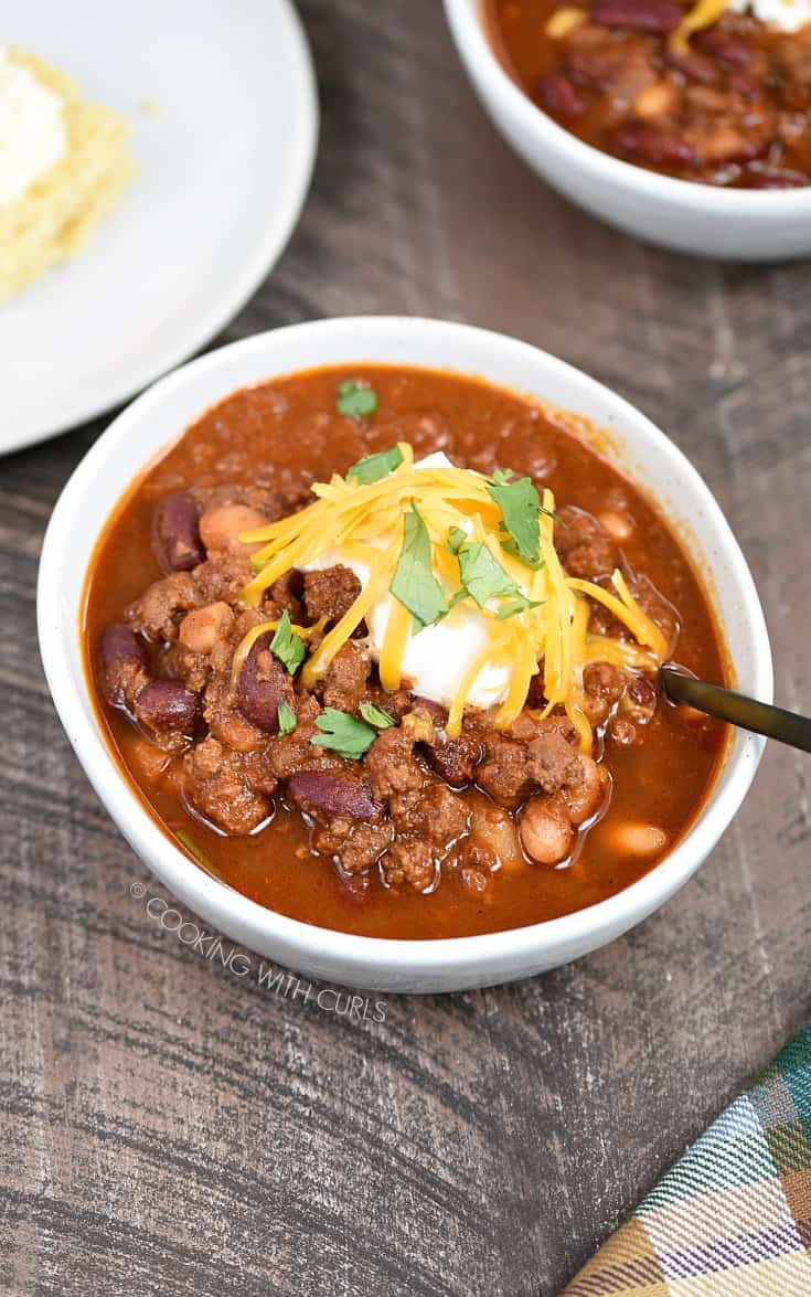 Looking down on a bowl of chili with a black spoon sticking out the side of the bowl and topped with sour cream, shredded cheddar cheese and chopped cilantro leaves.