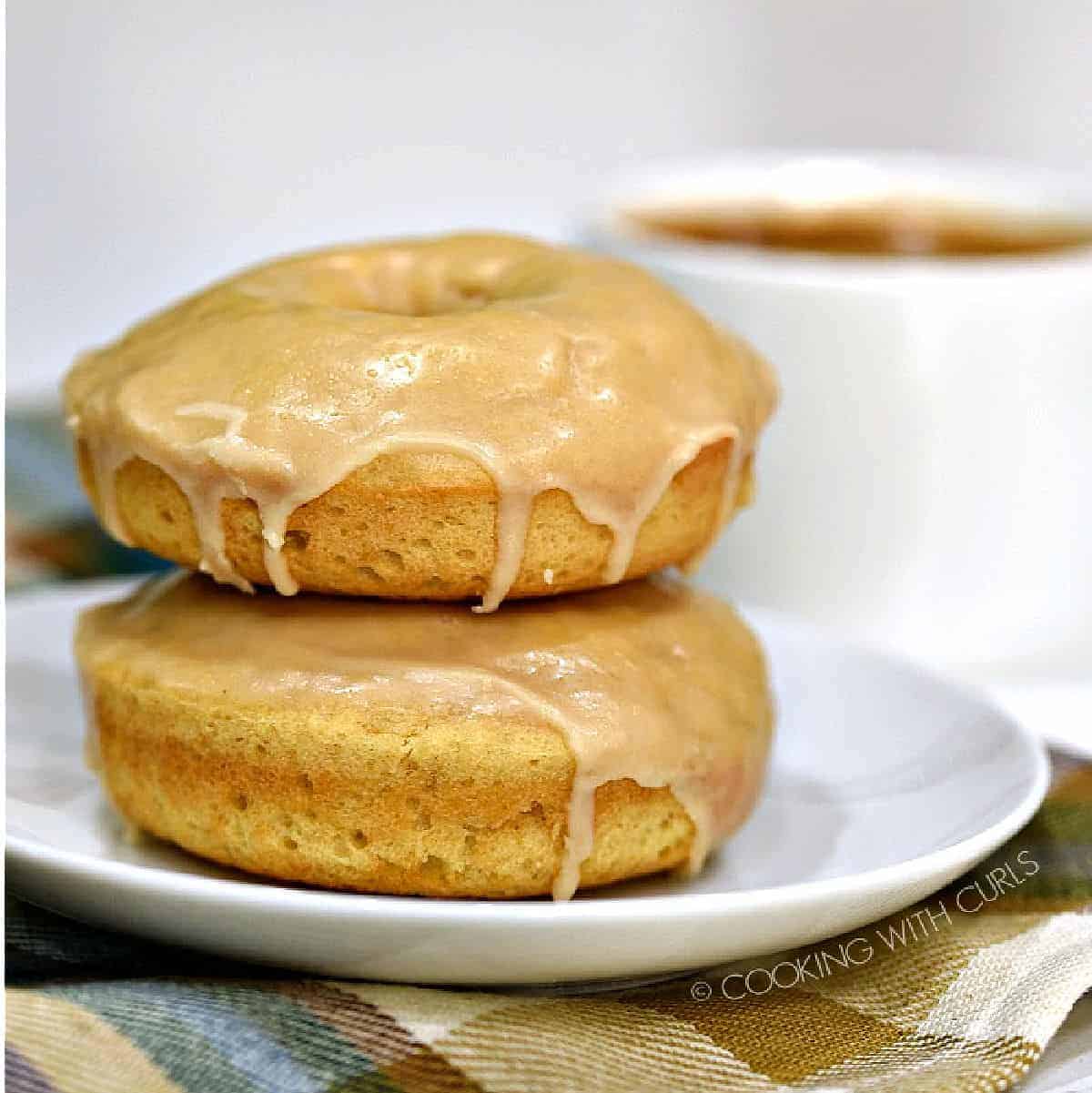 Two stacked maple glazed donuts on small white plate with a cup of coffee in background.