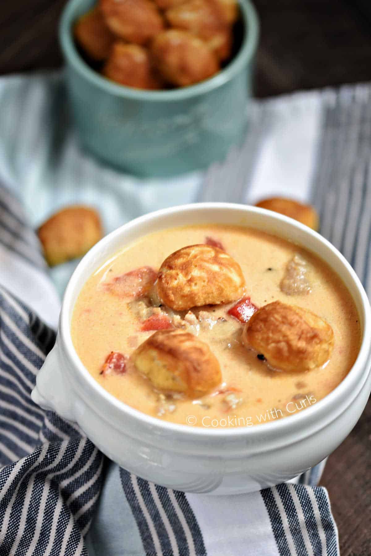 Brats and Beer Cheese Chowder topped with pretzel bites in a soup bowl with a bowl of pretzel bites in the background.