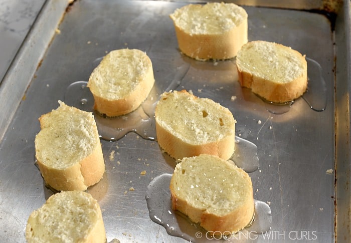 Seven bread slices on a baking sheet drizzled with olive oil.