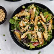 Two bowls of chicken shawarma salad with tahini dressing and pine nuts in the background.
