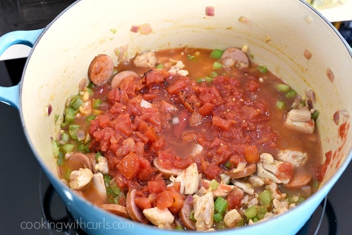 a large blue pot on a black cooktop filled with diced tomatoes, chicken chunks, bell pepper, onion, sausage and chicken stock