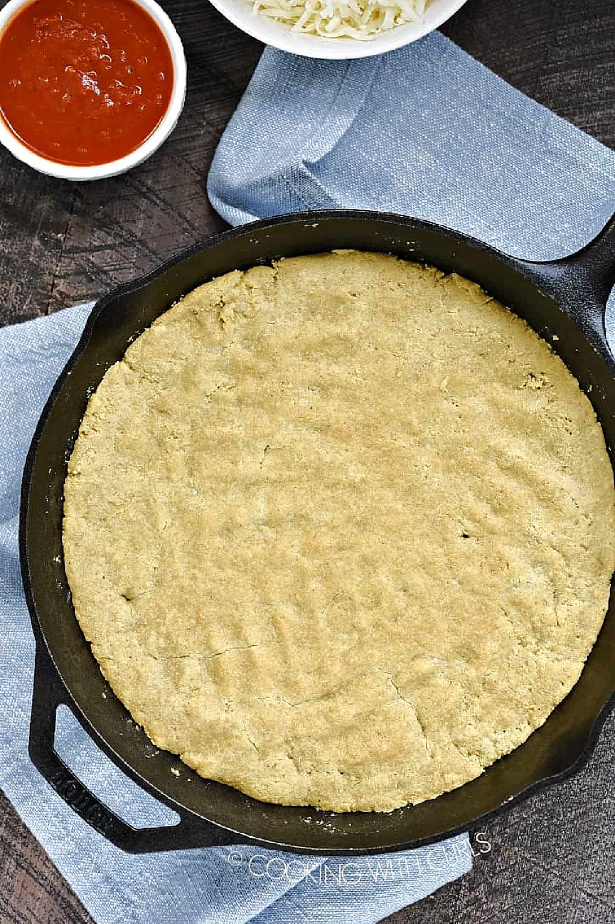 Baked Keto Pizza Crust in a cast iron skillet with bowls of sauce and shredded cheese in the background. 