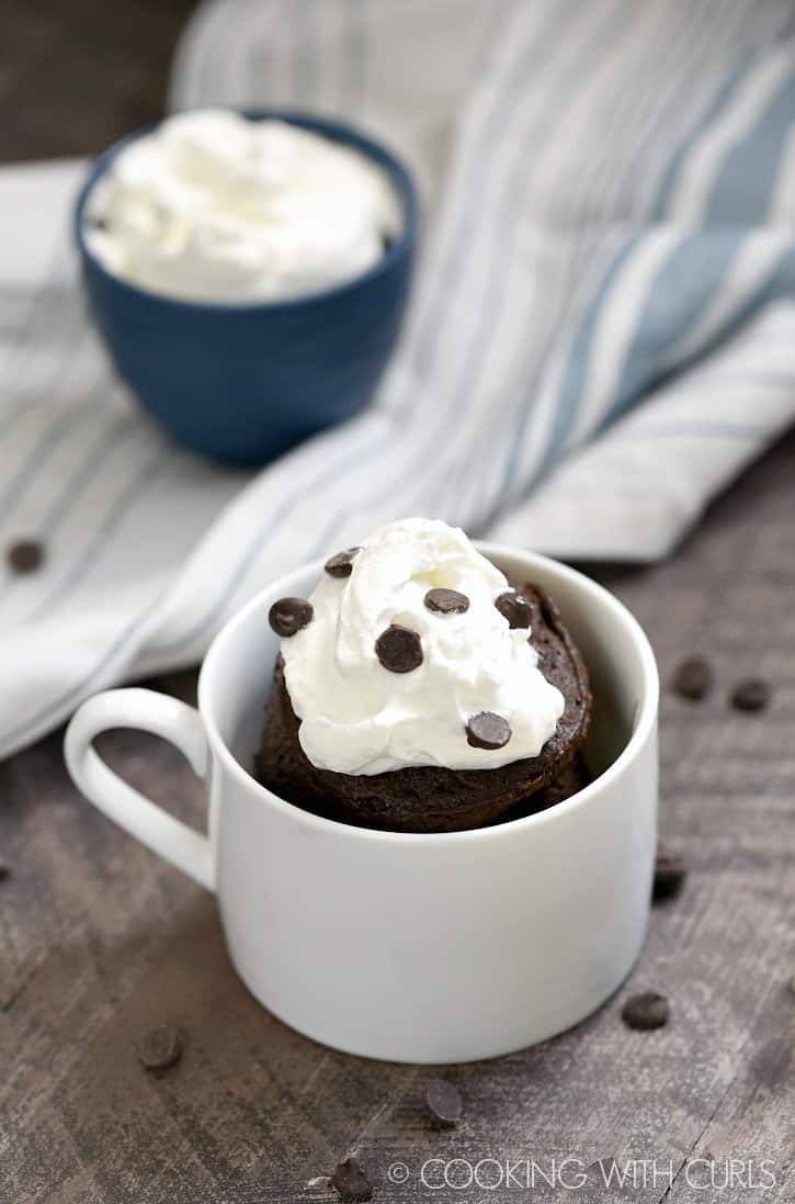 chocolate mug cake topped with whipped cream and mini chocolate chips in a small white mug with a small blue bowl of whipped cream in the background