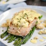 grilled salmon on a bed of asparagus topped with lump crab and bernaise sauce on an off-white plate with a second plate in the background