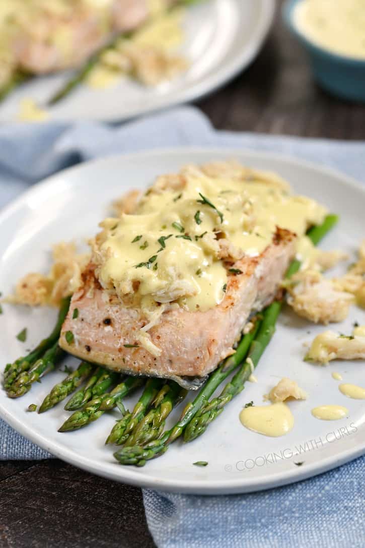 grilled salmon on a bed of asparagus topped with lump crab and béarnaise sauce on an off-white plate with a second plate in the background.