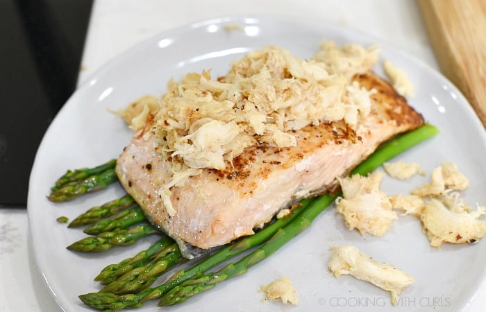 Salmon filet topped with lump crab on a bed of asparagus sitting on an off-white plate. 