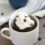 a whipped cream topped chocolate cake in a small white mug with mini chocolate sprinkles and a small blue bowl of whipped cream in the background sitting on a blue and white striped napkin