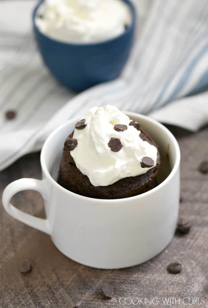 a whipped cream topped chocolate cake in a small white mug with mini chocolate sprinkles and a small blue bowl of whipped cream in the background sitting on a blue and white striped napkin