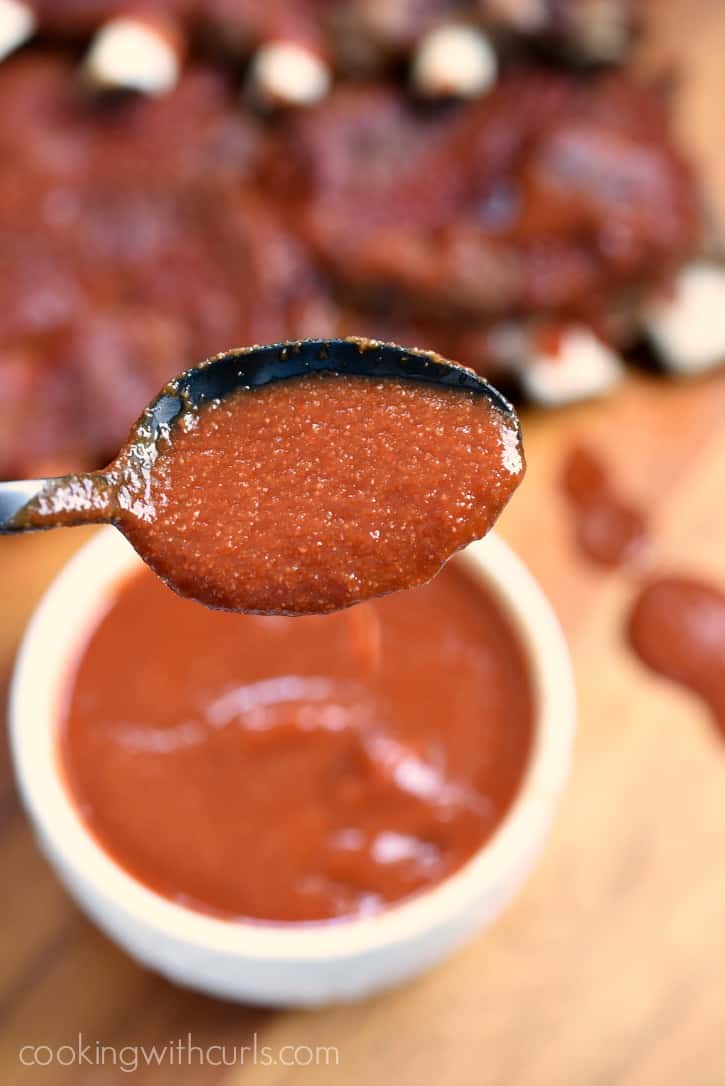 black spoon filled with barbecue sauce hovering over a small white bowl full of sauce with ribs in the background