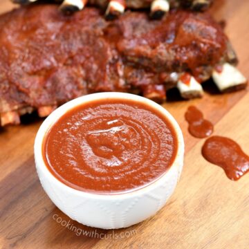 small white bowl sitting on a wooden board filled with sugar free bbq sauce with bbq ribs in the background