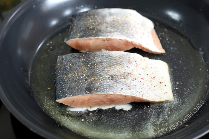 two skin-on salmon filets cooking in a non-stick skillet with butter and oil. 