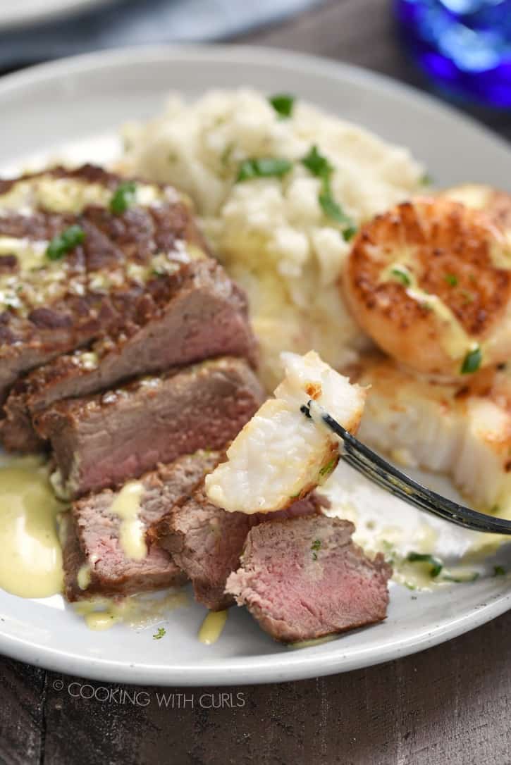 Sliced steak, mashed cauliflower, seared scallops and a piece of scallop on a fork hovering over the white plate