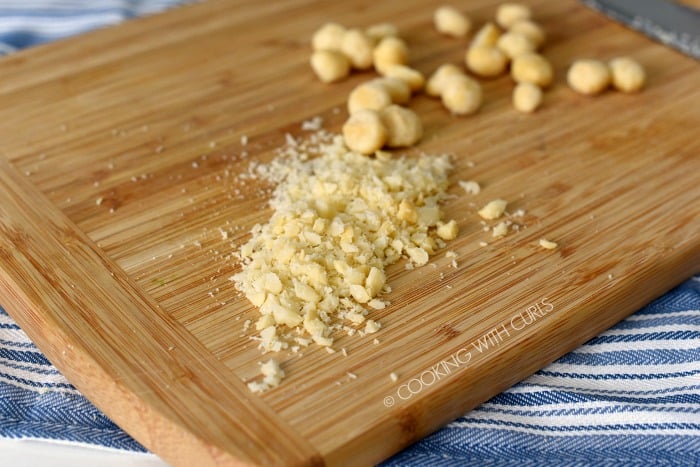 Chopped macadamia nuts on a wooden cutting board with whole nuts in the background