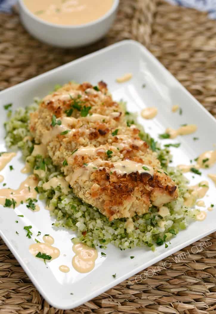 Macadamia Nut Crusted Mahi-Mahi drizzled with Sweet Chili Lime Butter Sauce served on a bed of riced cauliflower and broccoli on a square white plate with a bowl of sauce in the background