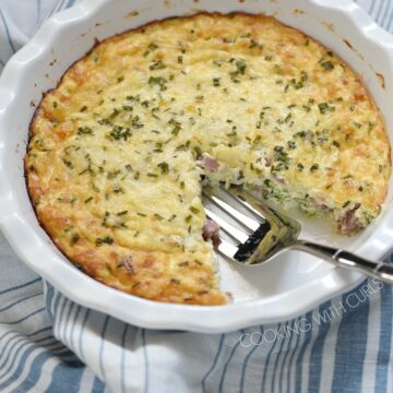 a white pie plate on a blue and white striped napkin, with one slice of crustless ham and cheese quiche removed and a serving fork in the void