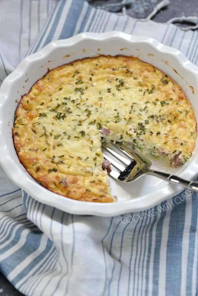 a white pie plate on a blue and white striped napkin, with one slice of crustless ham and cheese quiche removed and a serving fork in the void