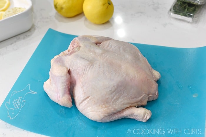 a raw, whole chicken laying on a blue plastic cutting mat with a white baking dish, two lemons, and two packages of herbs in the background 