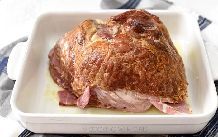 A spiral sliced ham placed cut side down in a white baking dish with pineapple juice, sitting on a blue and white striped towel. 