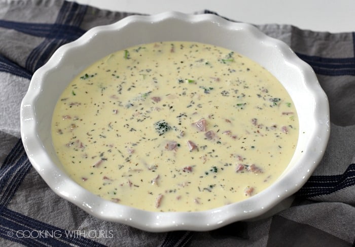 a white pie plate sitting on a blue and white striped dish towel filled with ham, broccoli and egg mixture for the crustless ham and cheese quiche