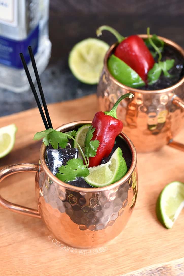 two copper mugs filled with ice cubes, lime juice and tequila, topped with lime wedges, cilantro sprig and a red chile pepper