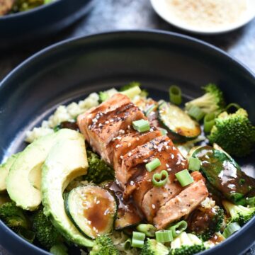 two blue bowls filled with vegetables and topped with salmon, teriyaki sauce, green onions and avocado. two small bowls with toasted sesame seeds and green onions are in the background