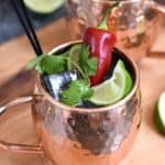 two copper mugs sitting on a wooden board filled with ice cubes, lime juice, tequila and ginger ale. They are topped off with a squished lime wedge, sprig of cilantro and a red chile pepper