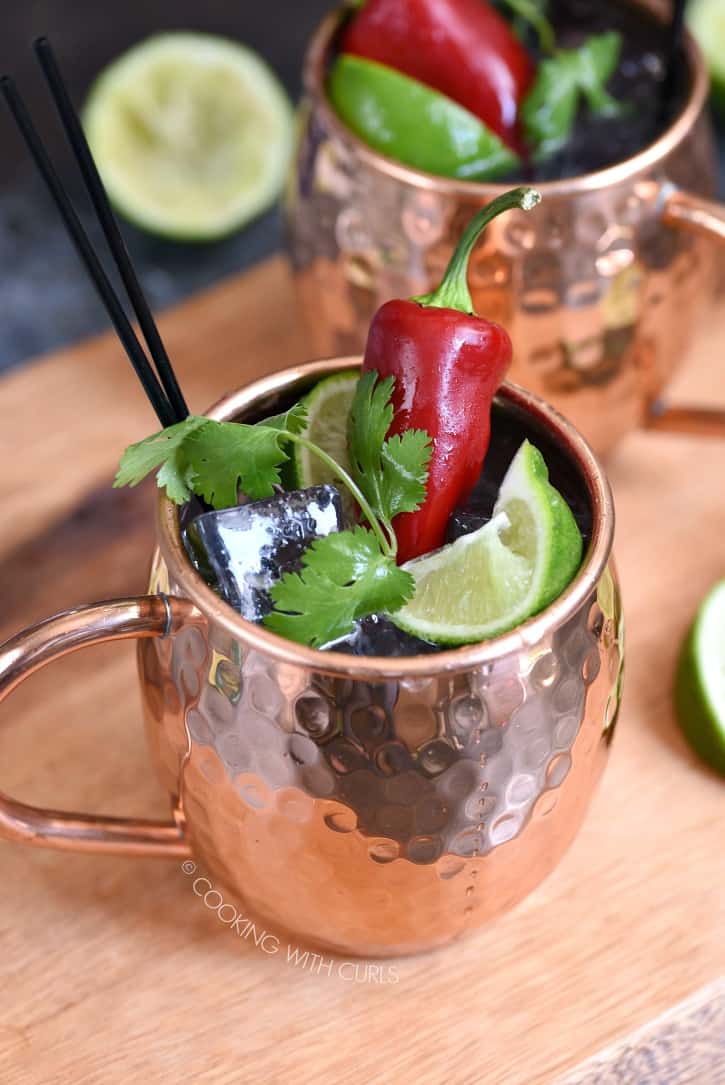 two copper mugs sitting on a wooden board filled with ice cubes, lime juice, tequila and ginger ale. They are topped off with a squished lime wedge, sprig of cilantro and a red chile pepper