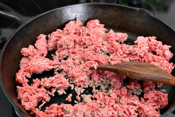 A cast iron skillet with ground beef and a wooden spatula in the bottom right hand corner 