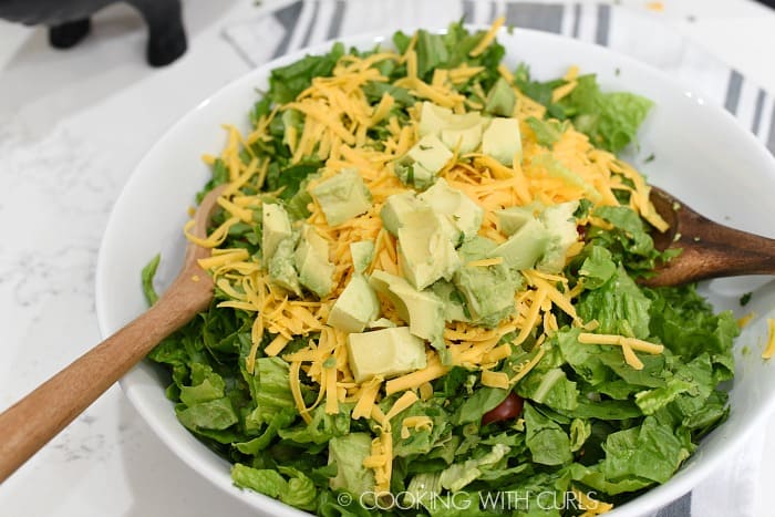 A large white bowl filled with lettuce, tomatoes, shredded cheddar cheese, avocado chunks and ground beef being tossed with two wooden spoons 