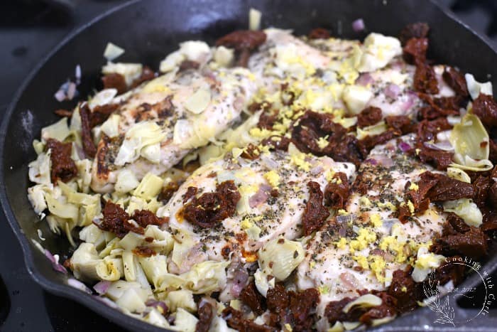 Four chicken breasts topped with sun-dried tomatoes, artichoke hearts and lemon zest in a cast iron skillet 