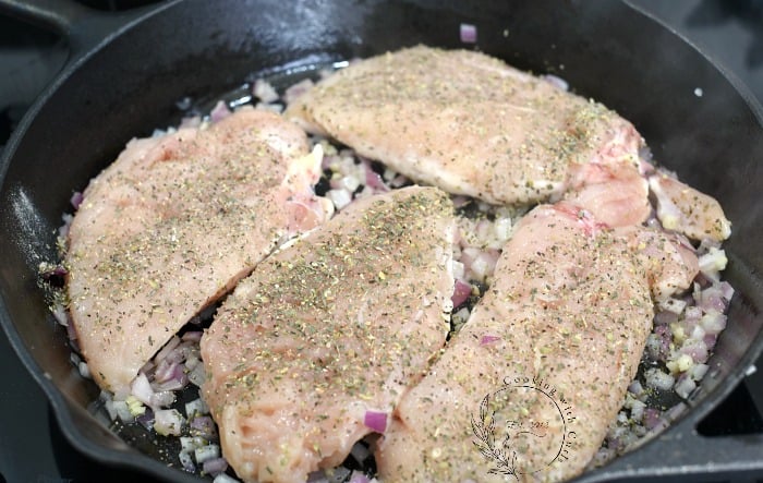 Four seasoned chicken breasts and diced red onion cooking in a cast iron skillet 