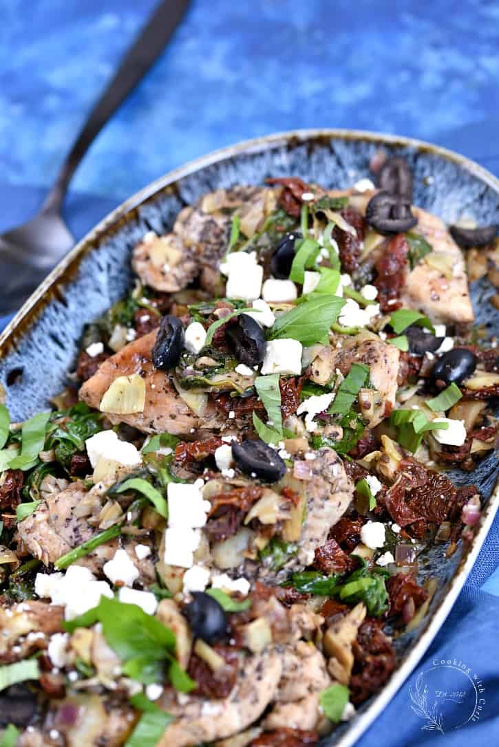 Chicken breasts topped with artichoke, tomato, olives, feta and basil in a blue bowl on a blue background