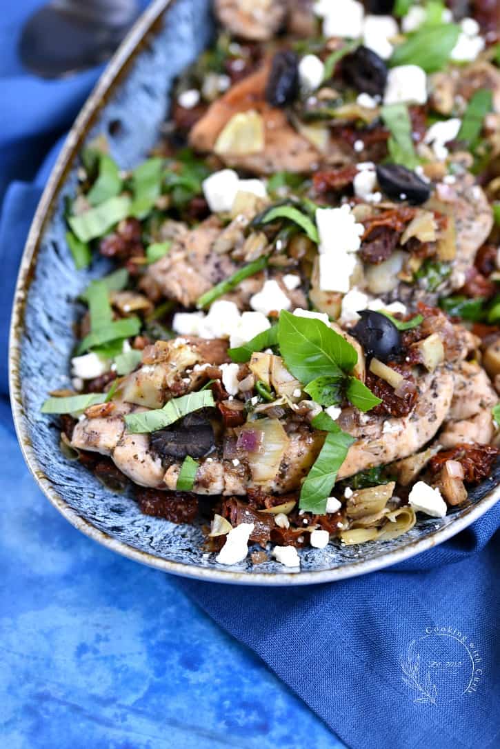 Chicken breasts topped with artichokes, tomato, olives, feta and basil leaves in a blue bowl sitting on a blue background